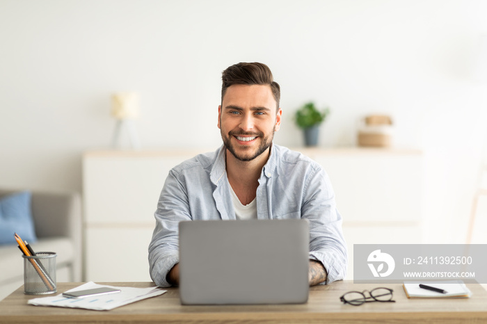 Successful freelancer. Happy man sitting at workplace with laptop computer, smiling and looking at camera