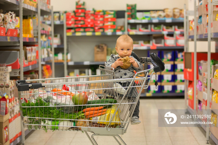 Toddler baby boy, sitting in a shopping cart in grocery store, smiling and eating bread