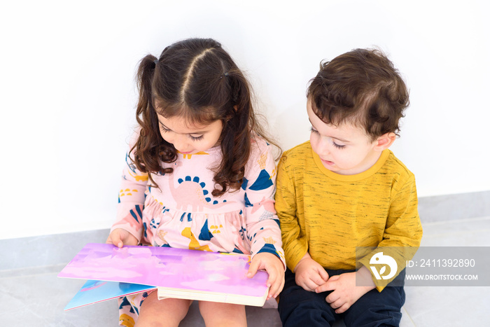 Two cute siblings reading book in home. Toddlers girl and boy having fun together. Happiness is in small things, slow living, education in home concept.