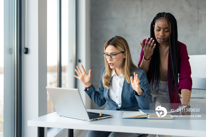 Two multiethnic business women in suit at office desk feeling disappointed, frustrated and sad over failure business plan, potential financial loss, looking at laptop screen in disbelief.