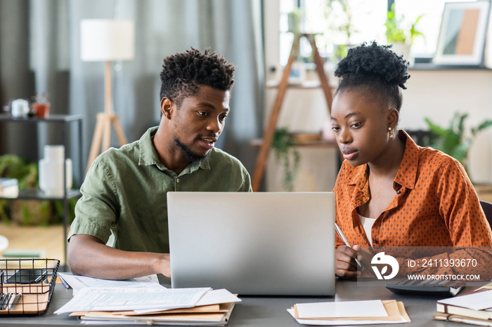 Young blackman sitting at table with bills and document and examining online tax form with girlfriend on laptop