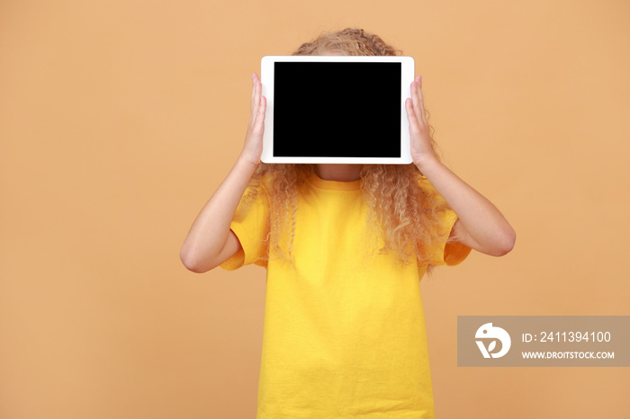 Amazed kid in hiding behind digital tablet with empty screen over yellow studio background. Excited schoolgirl showing pad, mockup, advertisement for cute application. New Educational App, Website