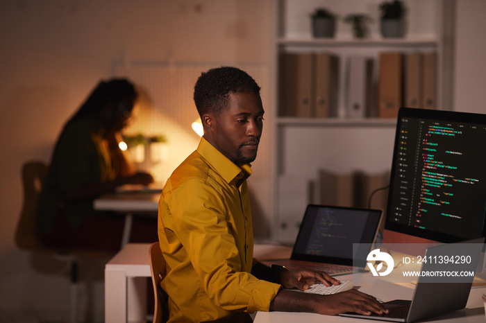 Portrait of African-American IT developer writing code on multiple computer screens while working late in office, copy space