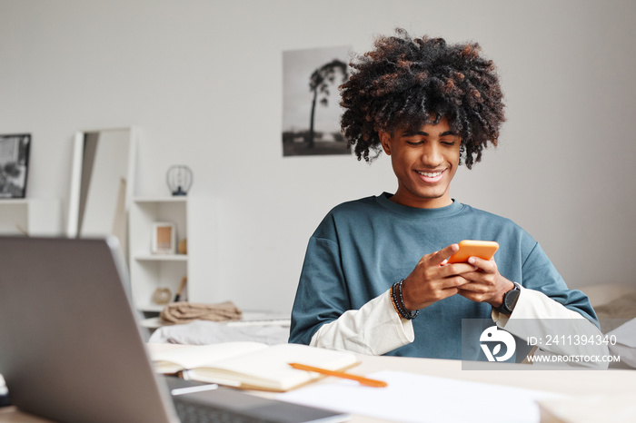 Portrait of African-American teenage boy using smartphone and smiling while studying at home or in college dorm, copy space