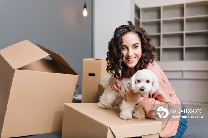 Moving to new apartment of young pretty woman with little dog. Chilling on bed surround carton boxes with pet, smiling to camera, expressing positivity