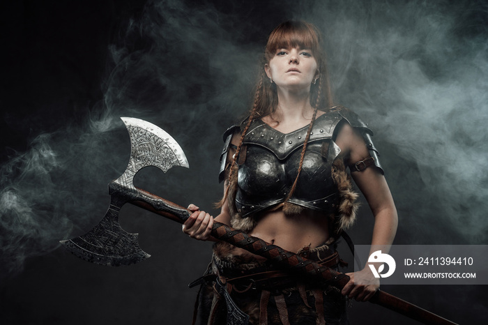 Barbaric beutiful woman from north armed with two handed axe and with brown hairs poses in dark foggy background.