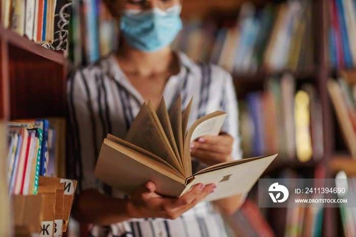 Closeup of attractive college girl standing in library with face mask on and reading a book. Studying during corona virus concept.