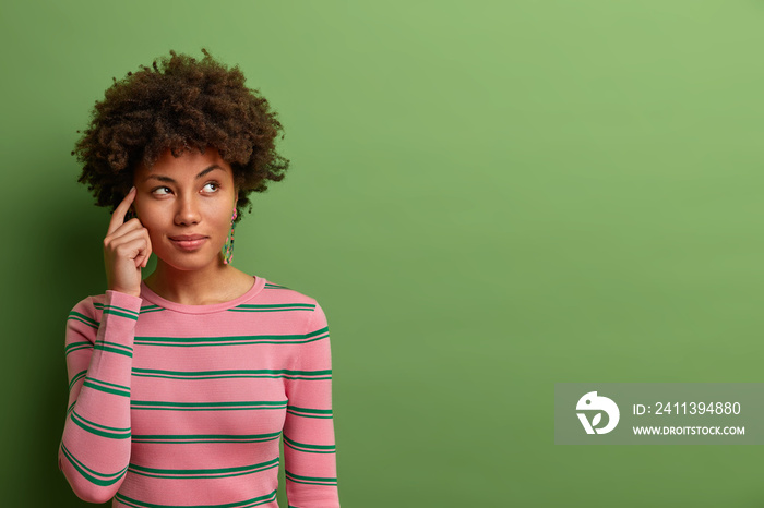 Half length shot of thoughtful dark skinned woman keeps fore finger on temple, thinks about offer or analyzes received information, dressed in striped jumper, poses against green background.