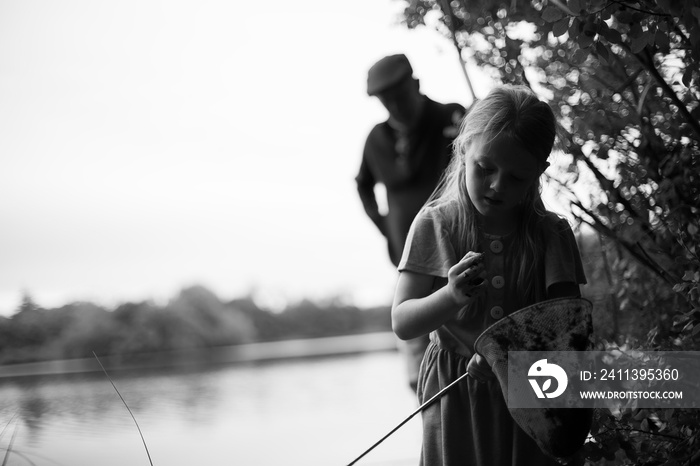 Young Caucasian female holding fishing net and examining something in another hand whilst her grandfather looks on in the background.