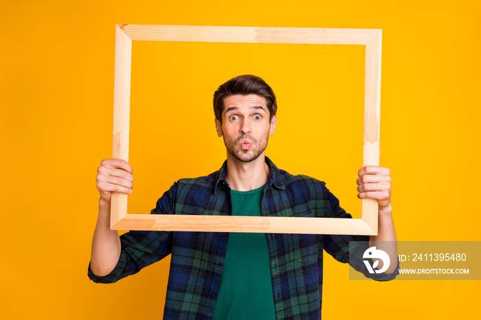 Photo of funny guy holding wooden picture frame looking through it sending air kiss wear casual plaid shirt isolated yellow color background