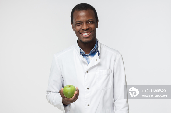 Indoor picture of young African American dentist standing in white uniform against white background with green apple in hand symbolizing necessity of taking care of teeth, smiling positively