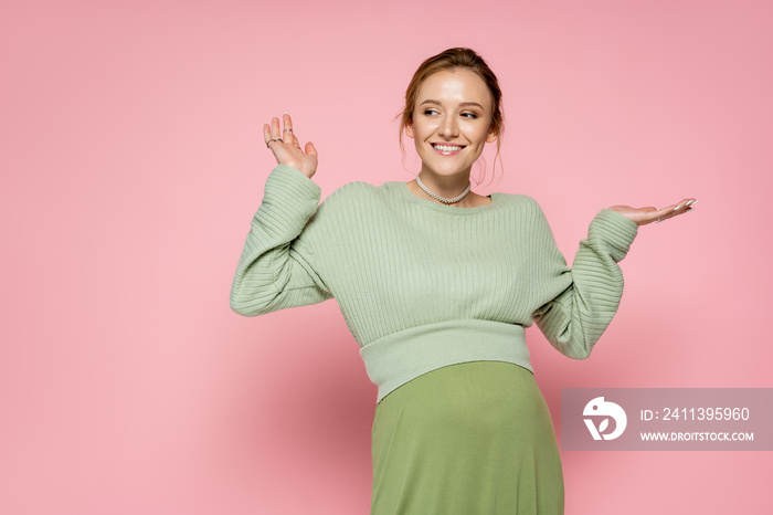 Positive pregnant woman in trendy green outfit pointing with hands on pink background.