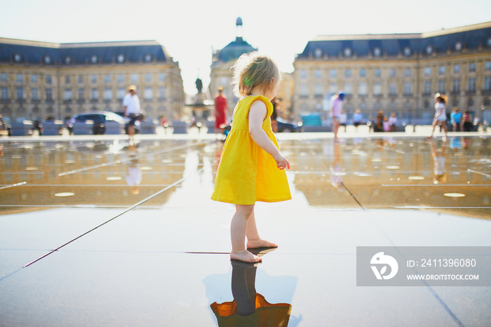 Adorable toddler girl in yellow dress having fun in famous outdoor water fountain in Bordeaux, France