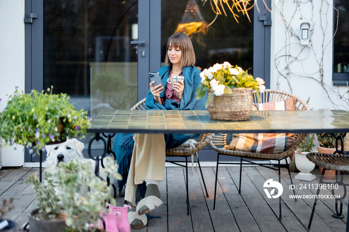 Young woman in plaid sitting relaxed by the table and enjoy drink at cozy home terrace with flowers in the evening