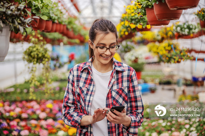 Young smiling female entrepreneur standing at hothouse and taking orders online. Hothouse interior.