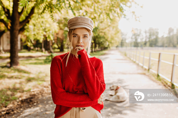 Beautiful young woman in red sweater and nice trendy hat looking thoughtful in autumn park. Attractive blonde in stylish clothes posing outdoor.