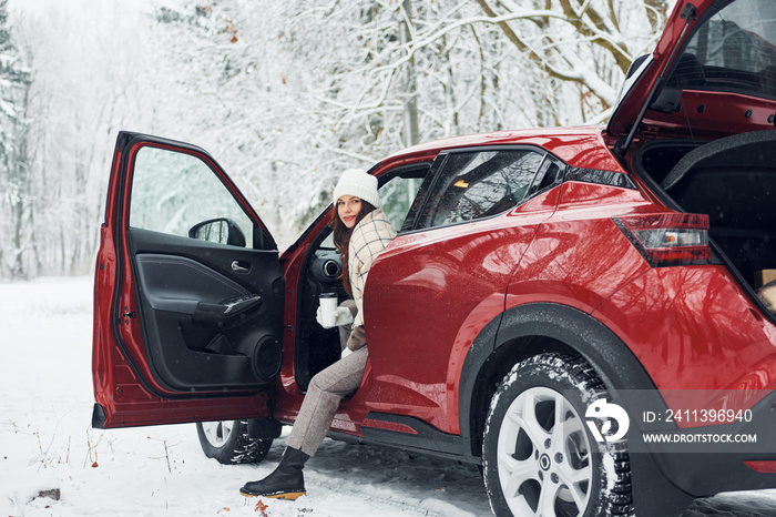 New car is in the forest. Beautiful young woman is outdoors near her red automobile at winter time