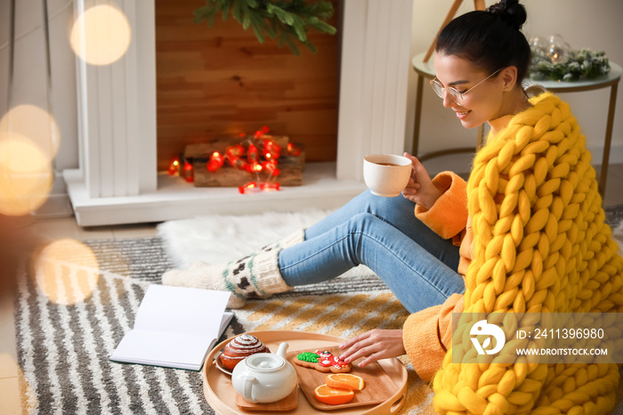 Beautiful young woman with warm plaid reading book and drinking tea at home