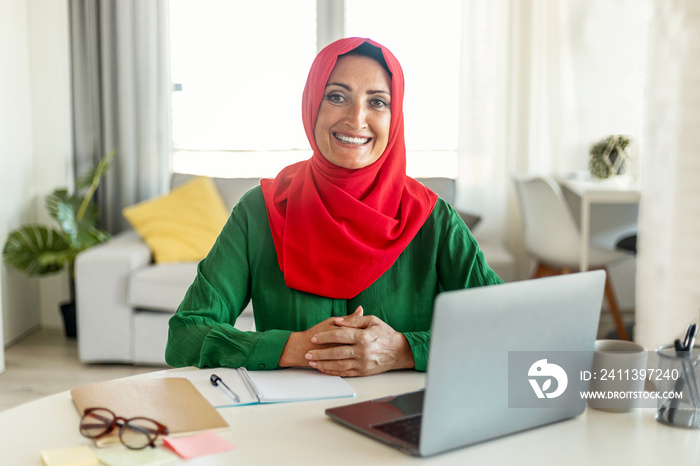 Portrait of happy mature woman in hijab using laptop pc, sitting at her desk and working online at home office