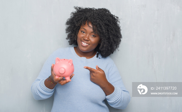 Young african american woman over grey grunge wall holding piggy bank very happy pointing with hand and finger