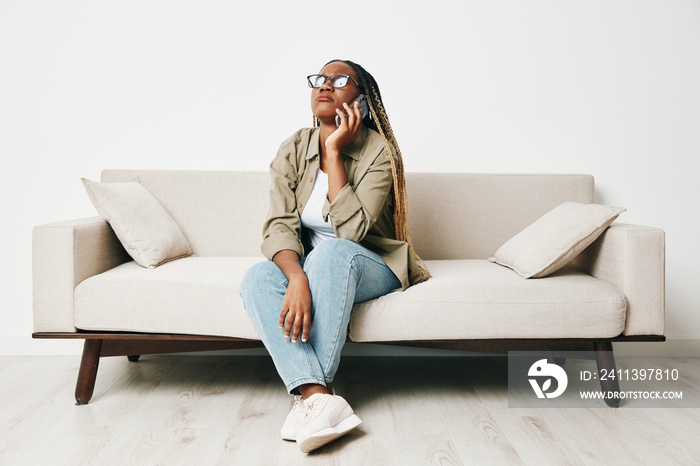 African American woman business freelancer working sitting on the couch at home in the phone, business calls and messages happiness smile, home clothes and eyeglasses, light interior background.