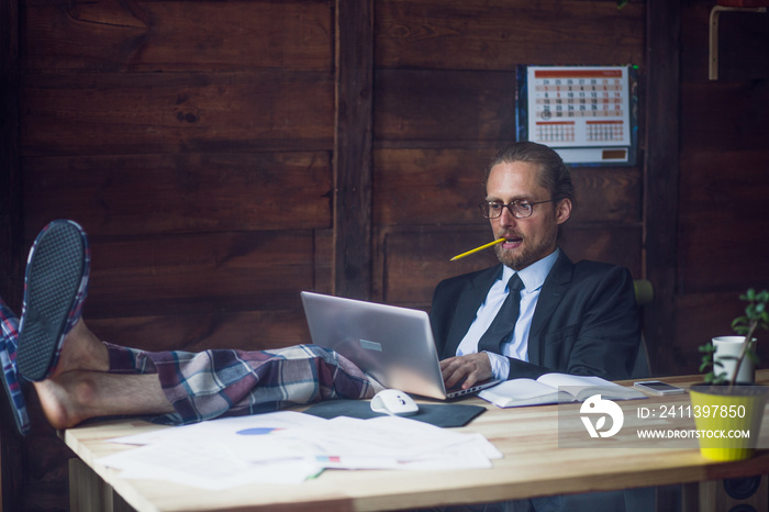 Freelance wearing business jacket, shirt and pajamas trousers. Man at home office holding legs on wooden desk while working with laptop. Toned image.