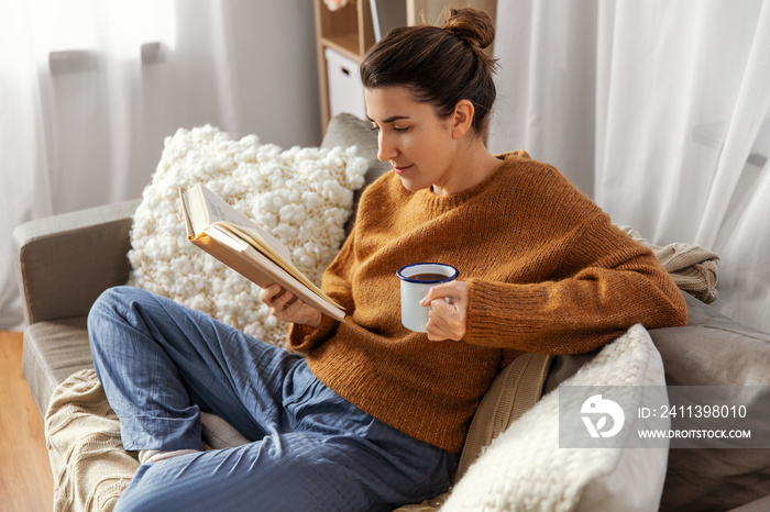 people and leisure concept - young woman drinking coffee and reading book sitting on sofa at home