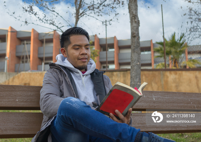 close up shot of a hispanic young man reading a book sitting on a park bench