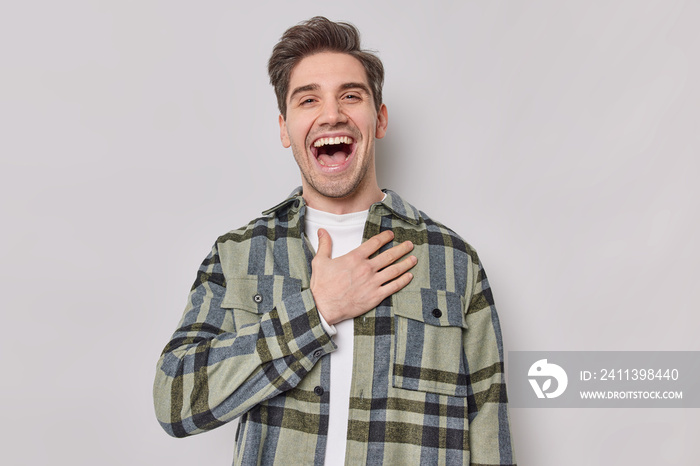 Optimistic glad handsome European man keeps hand on chest laughs joyfully hears funny story dressed in checkered shirt expresses positive emotions isolated over grey background. Happiness concept