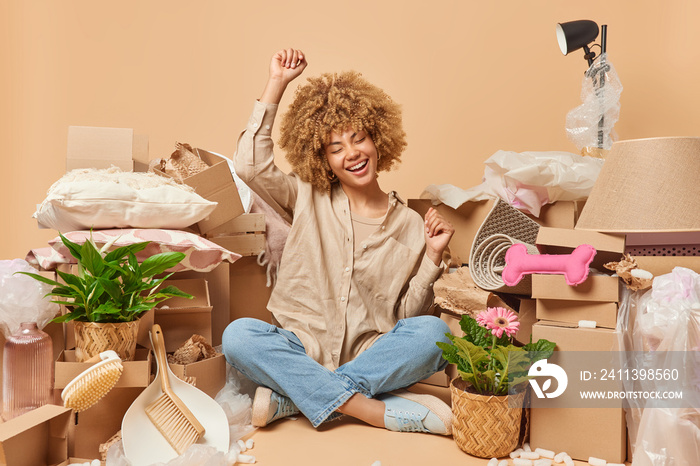 Optimistic curly haired woman wears shirt and jeans shakes arms sits crossed legs celebrates moving day collects personal belongings cardboard boxes isolated over beige background. Relocation concept