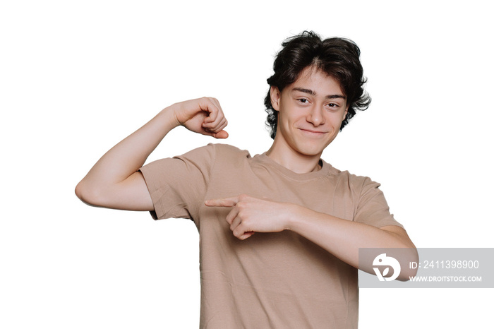 Handsome caucasian schoolboy in beige t-shirt bending arm demonstrates biceps pointing at it by index finger of another hand stands against transparent background. Cheerful Italian teen shows strength