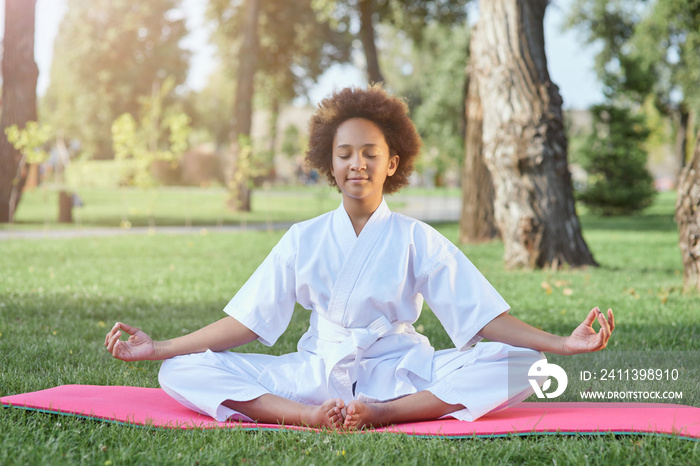 Cute Afro American girl in karate kimono meditating outdoors