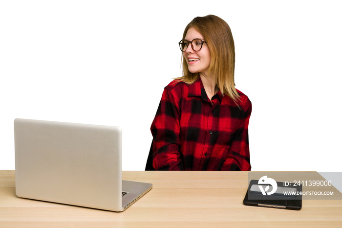 Young caucasian woman in a workplace working with a laptop isolated looks aside smiling, cheerful and pleasant.