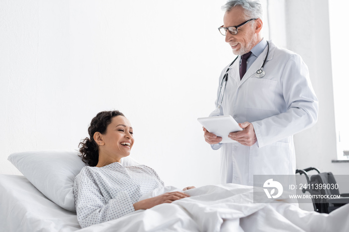 joyful african american woman lying in hospital bed near smiling doctor holding digital tablet