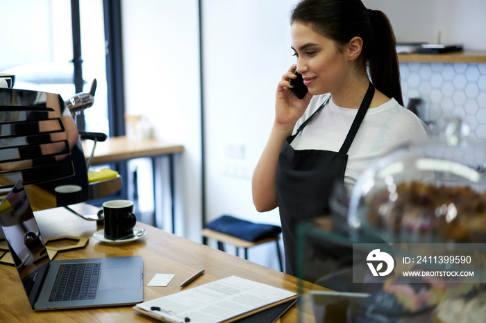 Professional brunette woman in apron having mobile phone conversation discussing business with delivery service operator, 20s skilled female manager of coffee shop ordering via smartphone call