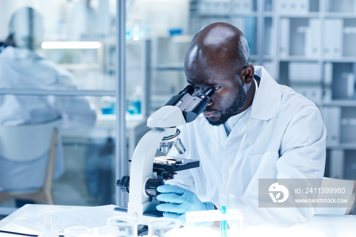 African scientist in lab coat concentrating on his work with microscope at table to examine the dna of virus