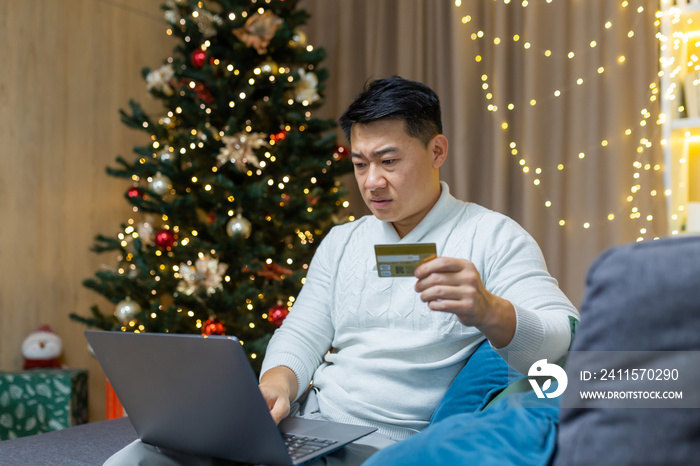 Confused and worried young handsome Asian man sitting at home and on sofa near Christmas tree. Holds