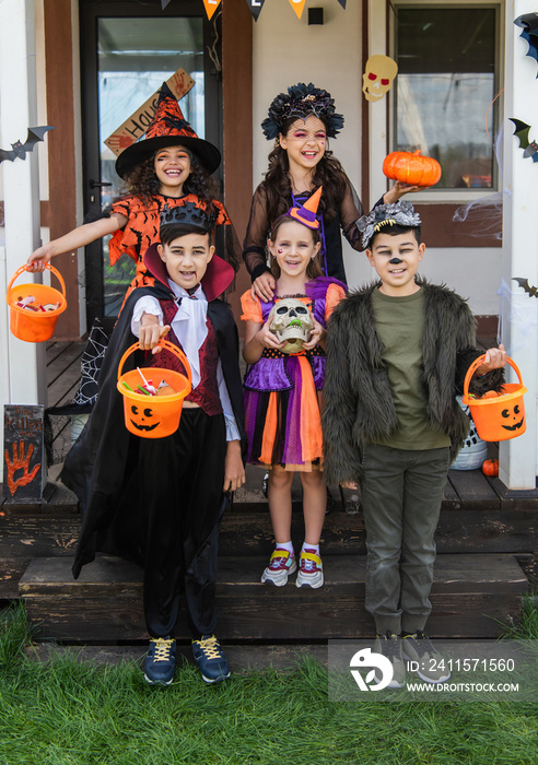 cheerful multiethnic friends in halloween costumes holding buckets with candies near decorated cotta