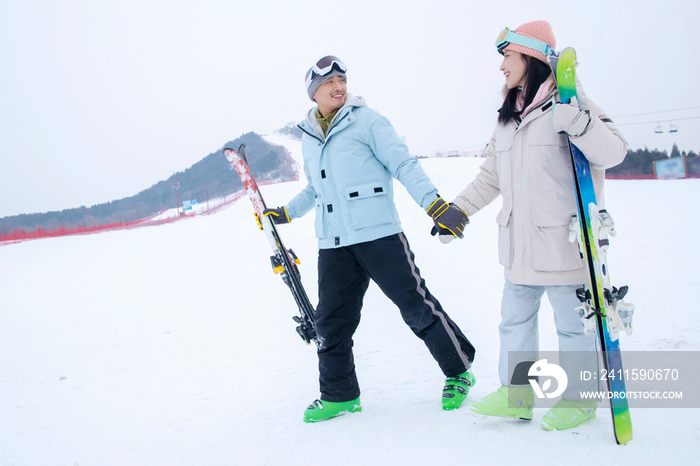 青年伴侣手牵手拿着滑雪板边走边聊