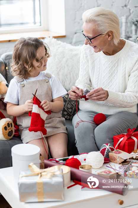 Grandmother and granddaughter doing craft toys and knitting near decorated Christmas new year tree. Cute little girl and attractive senior woman at home in the living room on the sofa having fun