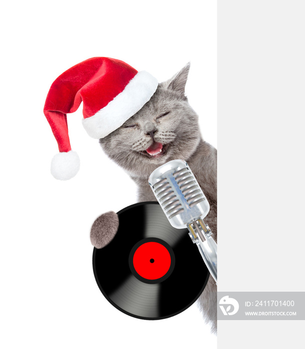 Kitten with retro microphone with vinyl record in red santa hat peeking from behind empty board. isolated on white background