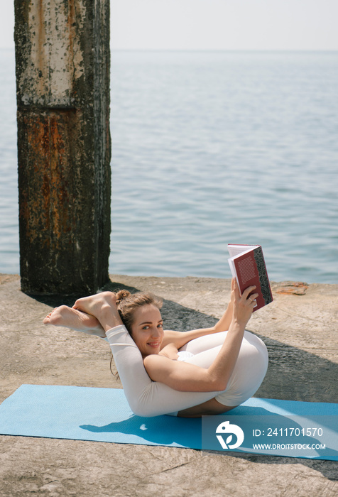Young woman reading a book while practicing yoga at seaside.  Woman doing yoga pose with legs behind her head