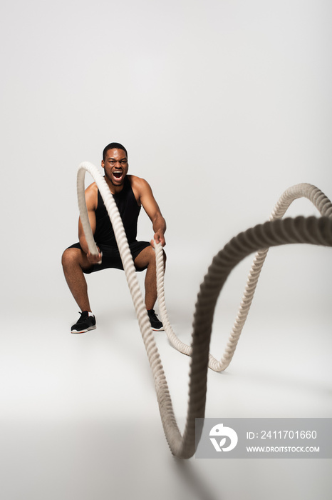 full length of african american sportsman screaming while exercising with battle ropes on grey.