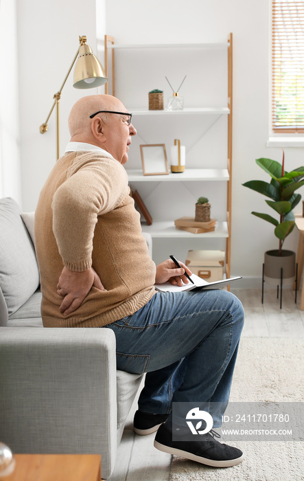 Senior man with notebook suffering from back pain on sofa at home