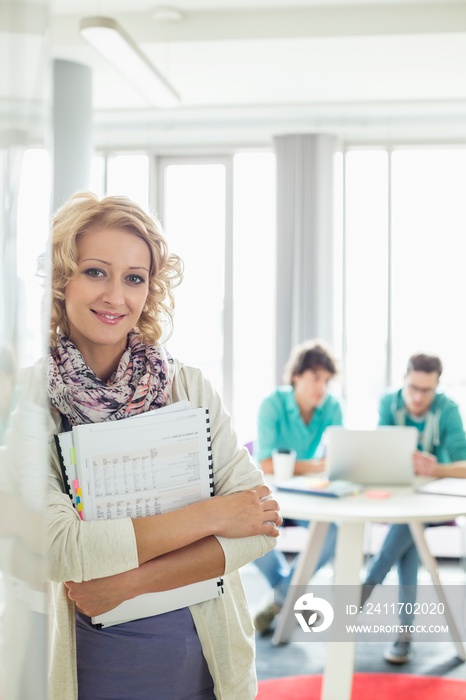 Portrait of beautiful businesswoman holding files with colleagues working in background at creative office