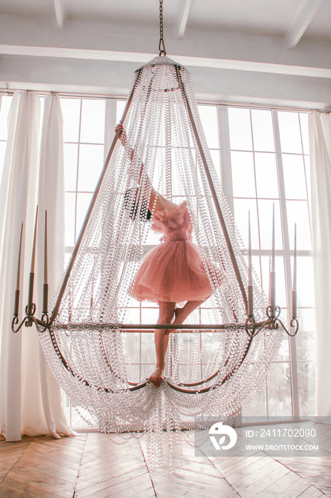 young girl with short black hair in pink princess dress is standing like ballerina on the crystal chandelier near huge white panoramic window background at sunny day. fashion concept, free space