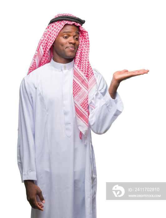 Young arabic african man wearing traditional keffiyeh over isolated background smiling cheerful presenting and pointing with palm of hand looking at the camera.