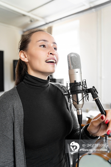 Young Asian or European cheerful brunette woman singing song to microphone alone in professional music studio or concervatory class. Radio host speaking to her listeners in broadcasting radio stream