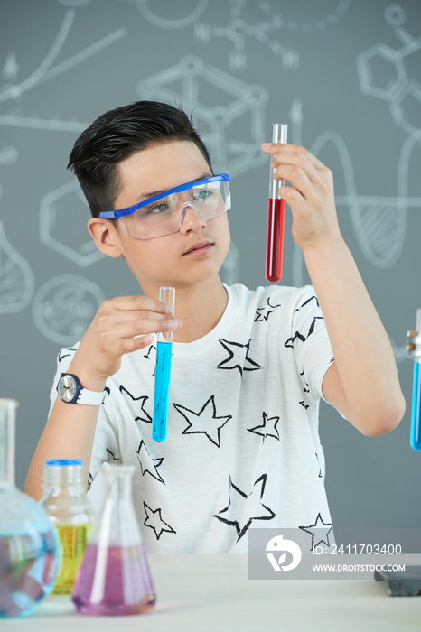 Curious pensive teen boy looking at test tubes with various reagents in his hands