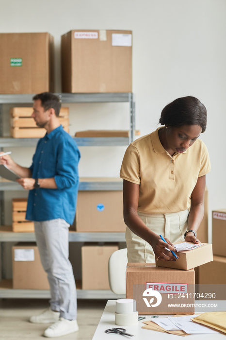 Vertical portrait of African-American woman packing boxes at warehouse while managing shipping service, focus on Fragile sticker